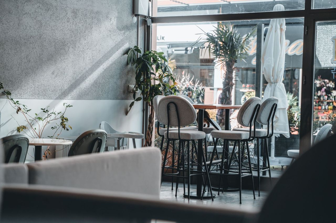 Bright café corner by a glass wall with high tables, upholstered chairs and plants.